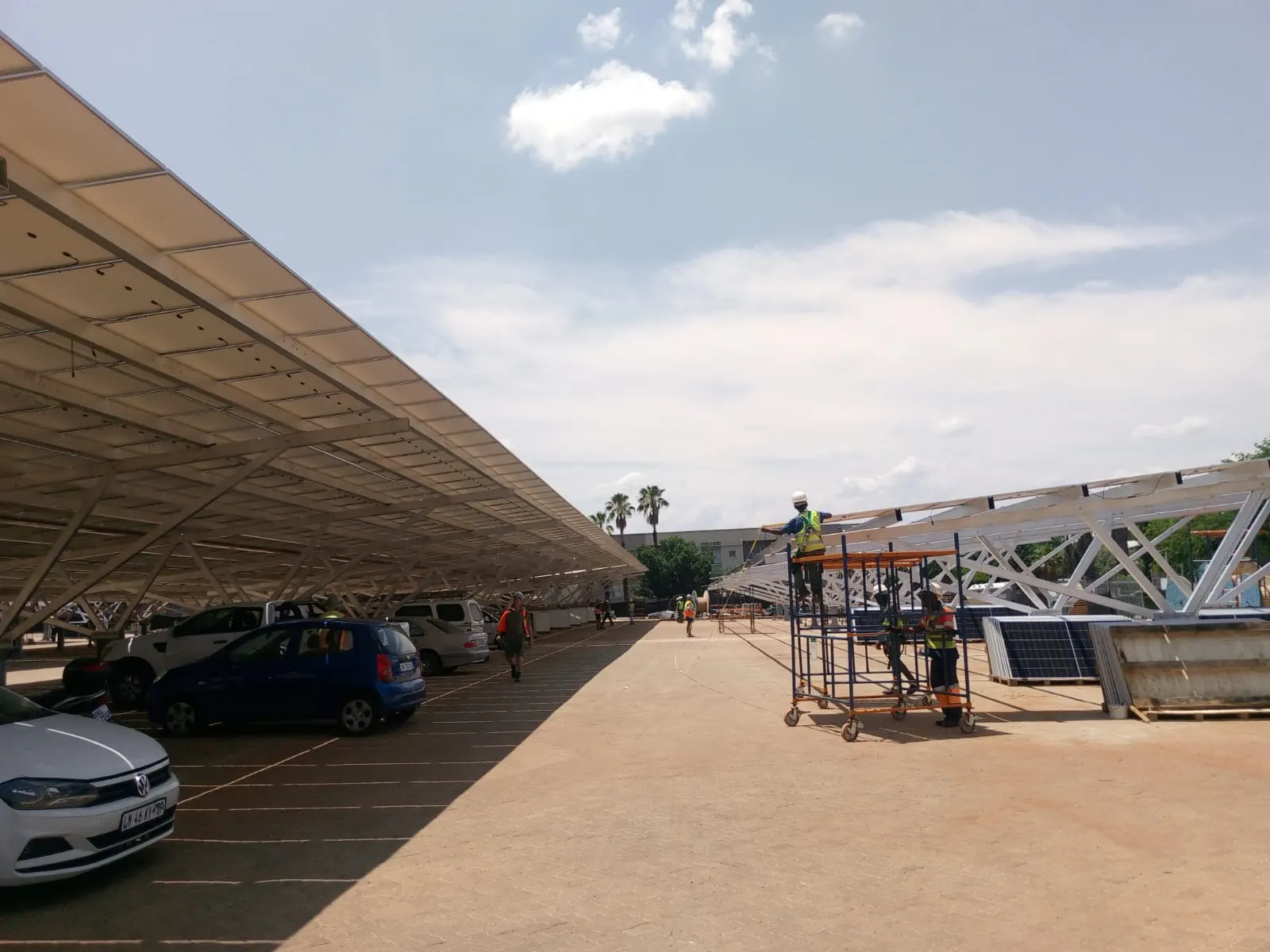 A completed solar carport structure spanning a parking area, with a second bay under construction beyond it.