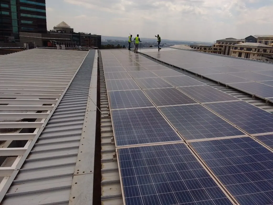 A wide rooftop solar installation on profiled sheeting, technicians working at the far end against a city skyline.