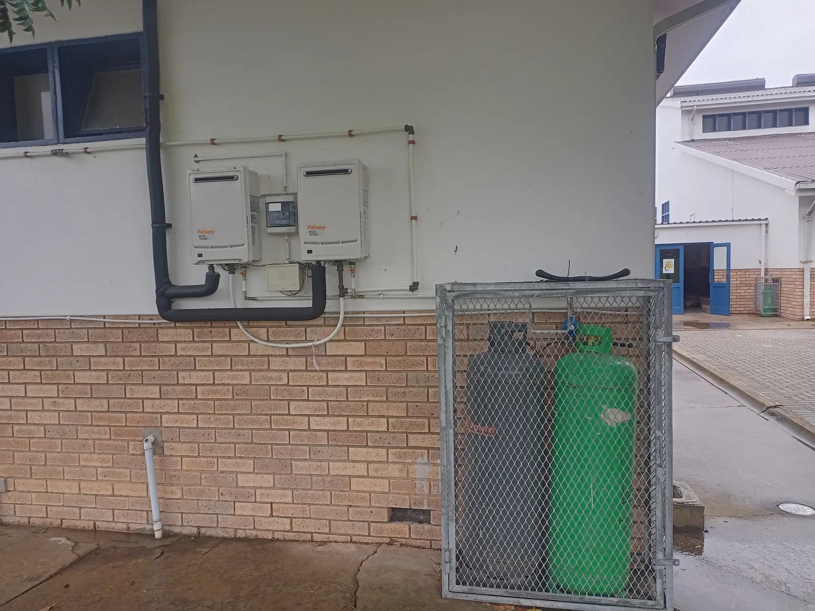 An external gas installation: a pair of instantaneous gas water heaters with the meter between them, hard-piped, and the supply cylinders secured in a lockable cage below.