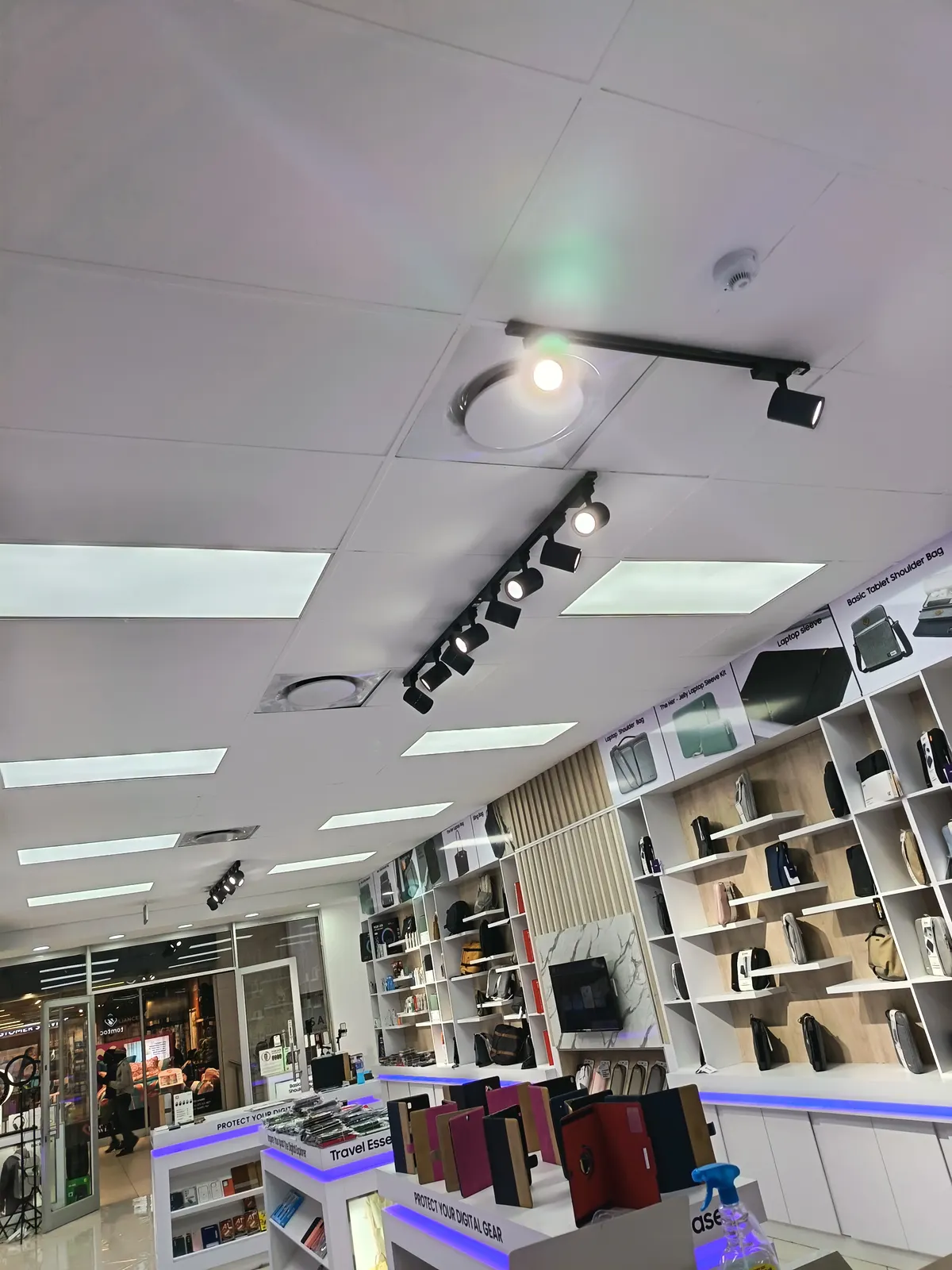 Track-mounted spotlights and recessed LED panels installed across a retail ceiling above display shelving.