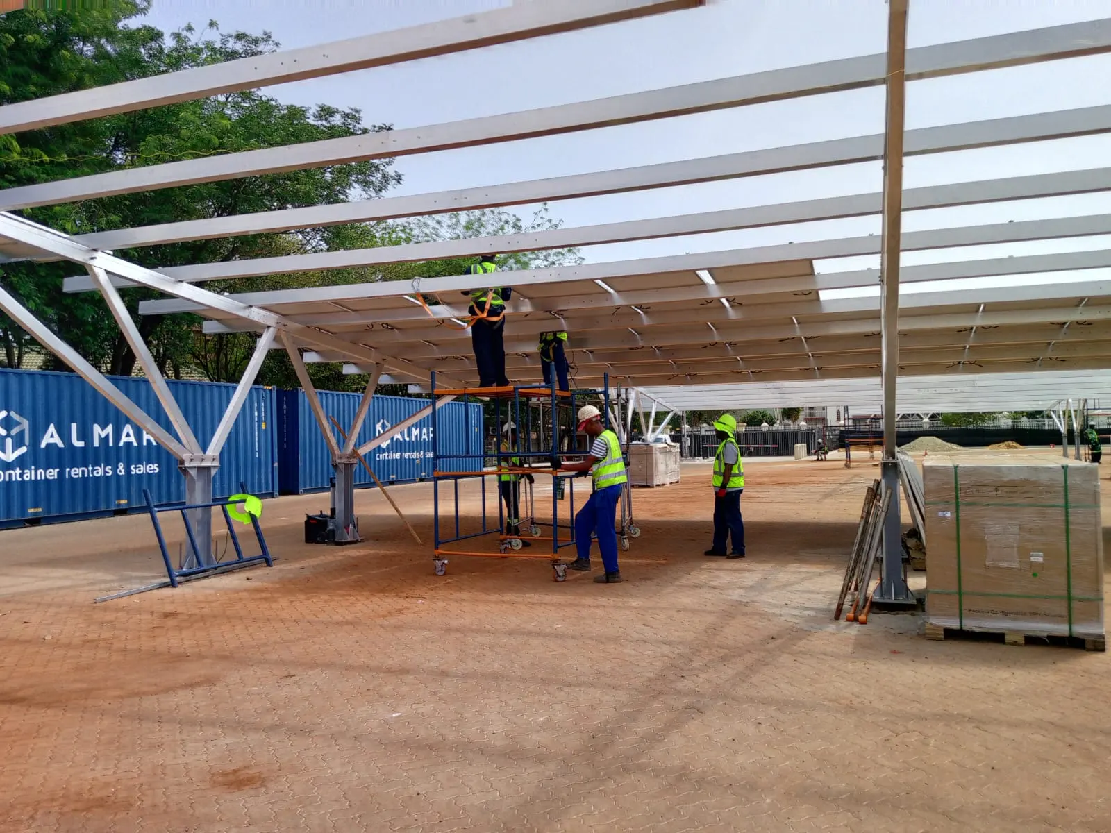 A crew erecting the steel frame of a solar carport, working from a mobile scaffold.