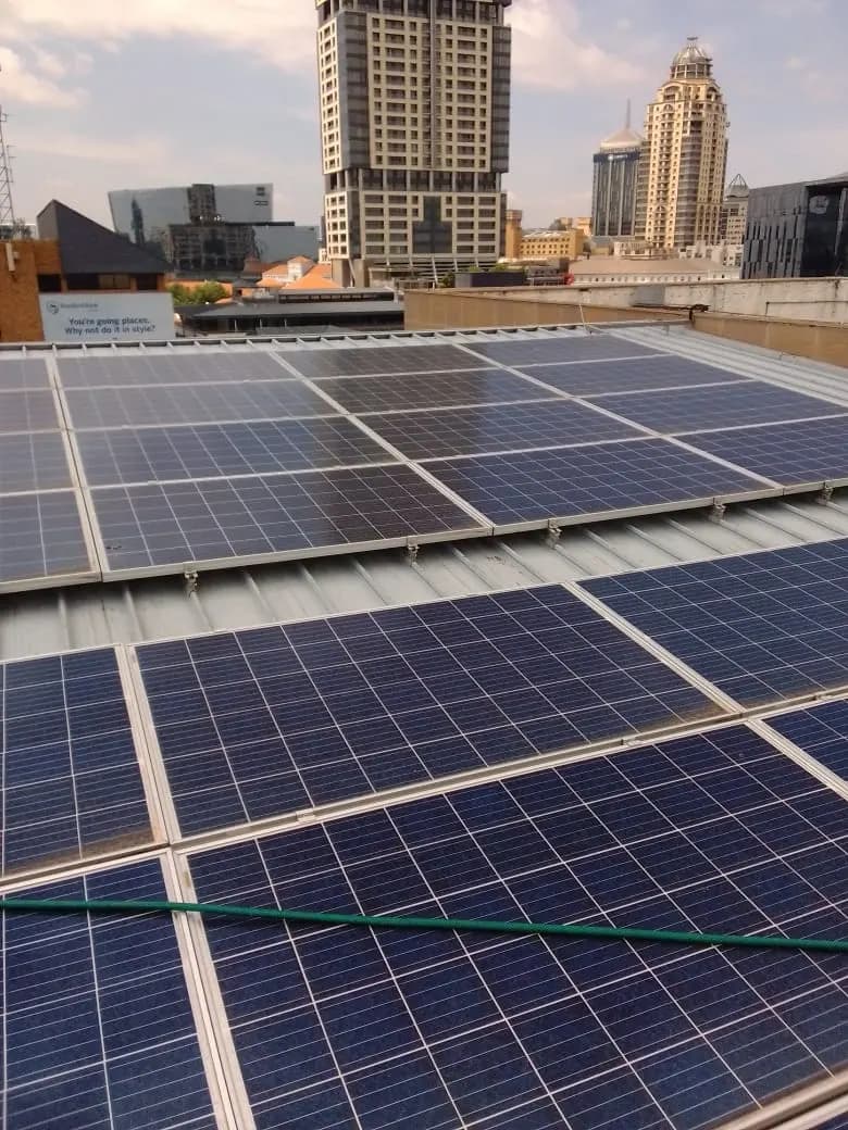 Rooftop solar panels mounted on rails above a sheet-metal roof, high-rise office towers in the background.