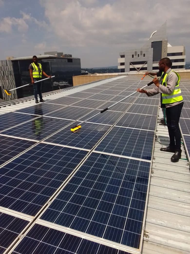 Two Powerline technicians in hi-vis vests washing down a commercial rooftop photovoltaic array, office towers behind them.