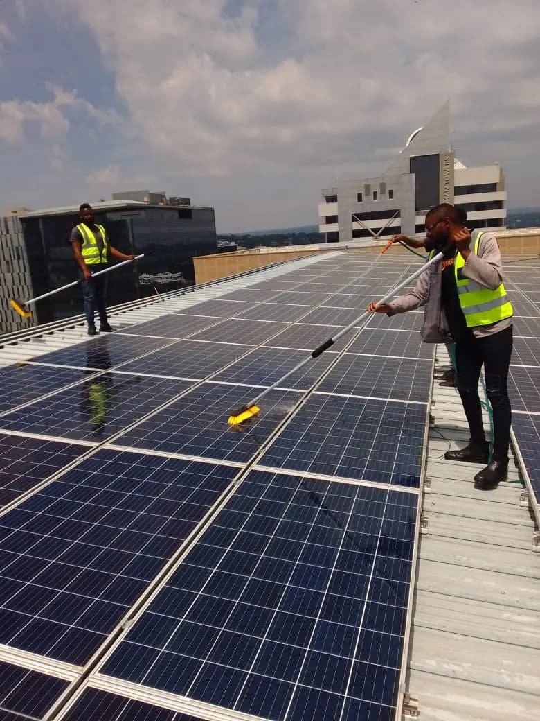 A technician working along a row of rooftop solar panels with a wash pole, a second technician at the far end of the array.